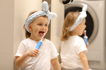 Adorible happy little girl in blue headbend holds toothbrush in bathroom. Beige background. Emotional impressed little girl opening mouth widely, having surprised facial expression. Hygiene concept.