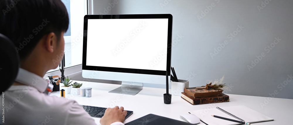 Poster rear view of young graphic designer sitting at his workspace in front of computer while working onli
