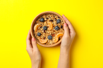 Female hands hold bowl with granola on yellow background, top view