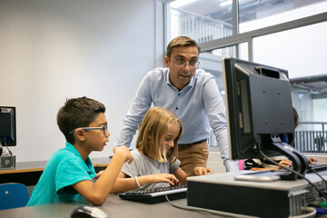 Happy kids sitting in front of computer monitor and learning IT. Caucasian teacher talking with multiethnic children about lesson and watching as they studying. Informatics and education concept