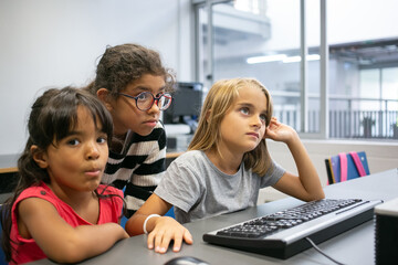 Pensive multiethnic girls studying together in computer class. Adorable little classmates in front of computer keyboard learning and thinking about task in classroom. Informatics and education concept