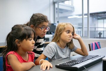 Three multiethnic girls sitting in classroom and studying together. Cute little classmates with computer keyboard learning and thinking about task in IT class. Informatics and education concept