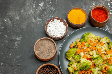 Half shot of healthy meal with brocoli and carrots on a black plate and spices on gray background