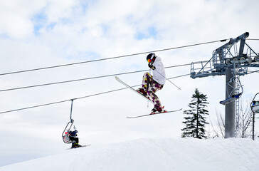 Fototapeta na wymiar Side view of man skier making jump in the air with cloudy sky and ski lifts on background. Male freerider on skis doing tricks while sliding down snow-covered slopes. Concept of extreme winter sports