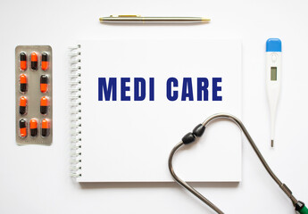 MEDI CARE is written in a notebook on a white table next to pills and a stethoscope.