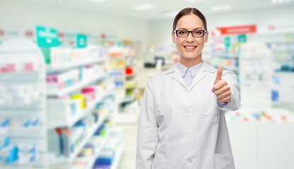 medicine, profession and healthcare concept - happy smiling female pharmacist or doctor in glasses and white coat showing thumbs up over pharmacy on background