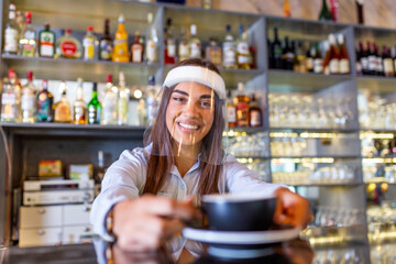 Beautiful female barista is holding a cup with hot coffee, looking at camera and wearing protective face shield while standing near the bar counter in cafe