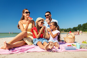 family, leisure and people concept - happy mother, father and two daughters having picnic on summer beach and eating watermelon