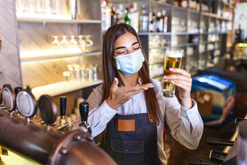 Beautiful female Bartender wearing protective face mask, serving a draft beer during coronavirus pandemic, shelves full of bottles with alcohol on the background