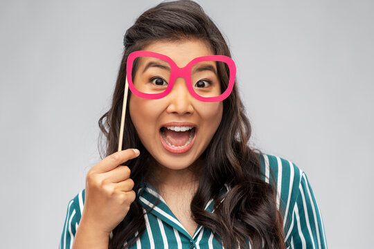 party props, photo booth and people concept - happy asian young woman with big glasses making faces over grey background