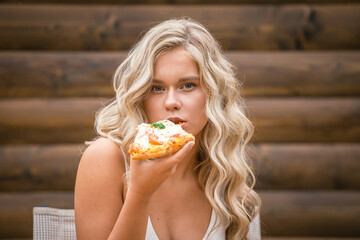 Beautiful girl with blond wavy hair in white clothes eating pizza on a wooden wall background