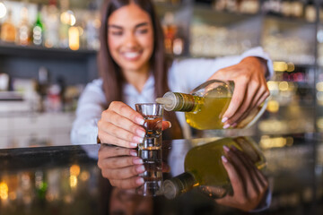 Beautiful female bartender is holding a shot glass with alcohol drink and a bottle in other hand, looking at camera and smiling while standing near the bar counter in cafe