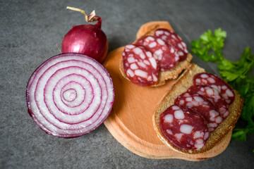 Black bread sandwich and salami sausage on wooden cutting board and cut purple onion