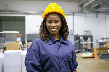 Cheerful African American female factory employee in hardhat and overall standing on plant floor,...