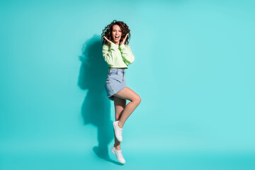 Full length body size photo of pretty girl jumping on one leg keeping hands near cheeks isolated on vivid teal color background