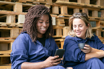 Fototapeta na wymiar Happy African American and Caucasian factory colleagues in overalls watching content on cellphone together while drinking coffee in warehouse. Labor or communication concept
