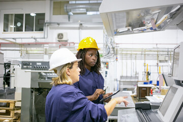 Female factory employees operating machine on plant floor together, using tablet, looking at...