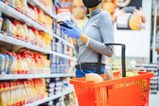 Unrecognizable Young Woman In Protective Gloves And Mask Holding Basket With Groceries And Smartphone While Shopping In Supermarket