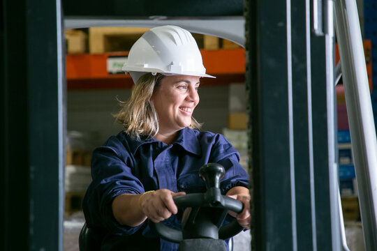 Happy Caucasian Female Logistic Worker In Hardhat Driving Forklift In Warehouse, Smiling, Looking Away. Side View. Female Labor Concept