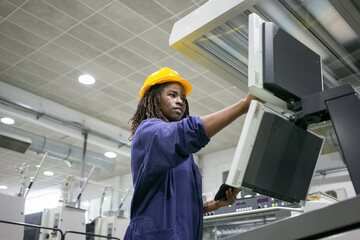 Focused confident black female factory worker operating industrial machine, touching control board, using tablet. Low angle, copy space. Production process or machinery concept