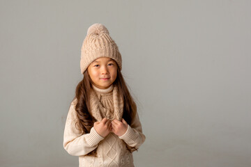 little girl of Asian appearance in a knitted sweater and a hat on a gray background