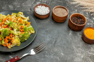 Top view of set of different spices in brown bowls and vegetable salad and fork on gray table