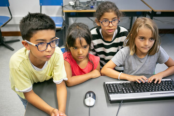 Multiethnic children sitting at desk during IT lesson and doing task. Thoughtful adorable kids...