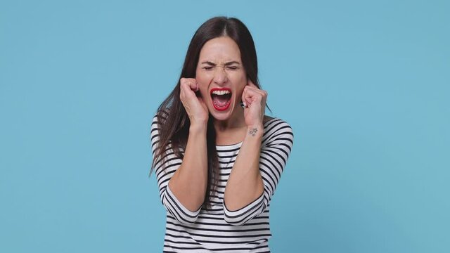 Crazy frustrated brunette young woman 20s wearing basic striped shirt posing isolated on blue background in studio. People lifestyle concept. Covering ears with fingers keeping eyes closed screaming