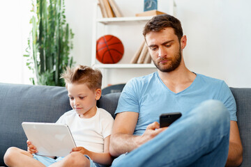 Little boy using digital tablet and his father using smartphone sit together on couch