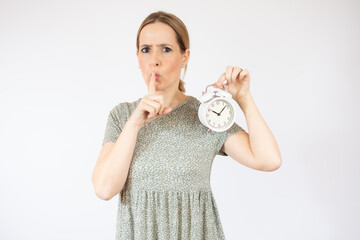 Portrait of beautiful and young woman standing over isolated white background holding an alarm clock and asking to be quiet with finger on lips. Silence and secret concept.
