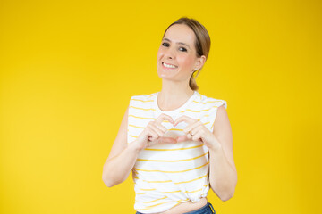 Young woman in casual t-shirt making heart figure over yellow background.