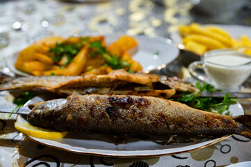 Grilled trout on white plate with lemon pieces on kitchen towel on wooden table.