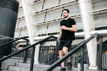 Athletic young man running down from stairs. Urban sport concept. Apartment block on background. Active living. Outdoor fitness. Man in black sportswear jogging at morning.