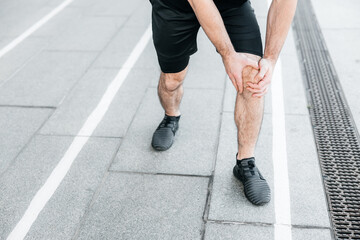Male hands holding aching knee. Strong man feeling pain in his foot during jogging outdoors. Close up. Male runner in black sneakers standing and feeling cramps in his left leg. Cut view.