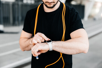 Athletic man with orange jumping rope around neck setting up his smart watch. Cut view of confitent male runner which records the results of the run on the device. Modern technologies concept.