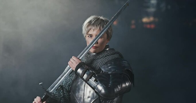 Dramatic shot of a woman in a metal medieval warrior armour with a sword, examining the blade, while standing in dark environment with smoke in background