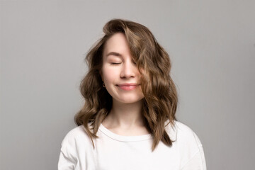 Obraz premium Portrait of happy smiling young woman with closed eyes, gray background, copy space. Beautiful caucasian girl with brown curly hairstyle. Cheerful, satisfied, dreamy expression face. Calmness, relax