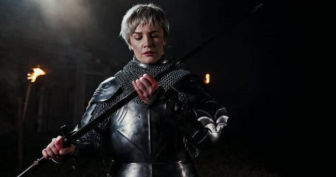 Adult woman with short haircut in Joan of Arc armor, standing in dark room with torches and examining her sword blades sharpness after the fight