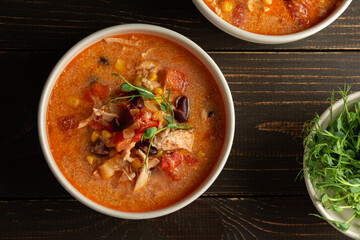 Spicy mexican soup with beans and corn, tomatoes, chicken breast and bean sprouts, bowl with soup and napkin on a dark wooden background, top view