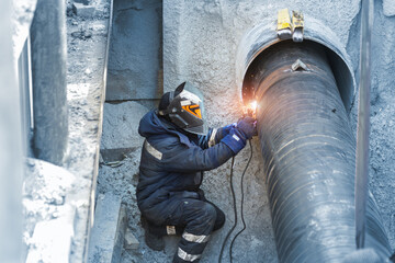 Professional welder welding water or gas steel pipeline in protective trench and mask at...