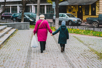 Grandma in pink jacket and white plastic bag walking with granddaughter in black jacket on the...
