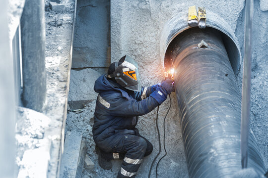 Professional welder welding water or gas steel pipeline in protective trench and mask at construction site pit. City underground utilitites renewal replacement. Emergency pipe break leak elimination
