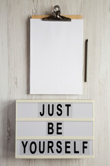 'Just be yourself' on a lightbox, clipboard with blank sheet of paper on a white wooden surface, top view. Flat lay, overhead, from above.