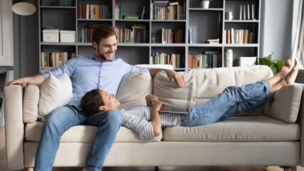 Wide banner view of happy young Caucasian couple relax on couch in living room using laptop browsing internet together. Smiling man and woman rest on sofa at home use computer shopping online.