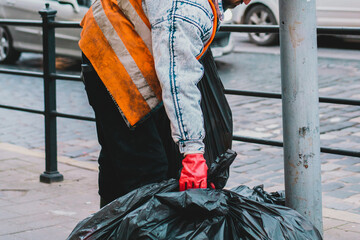 Janitor in red gloves and yellow uniform on the city’s street takes a few big black bags of...