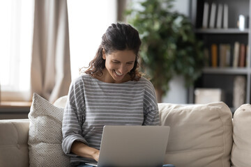 Naklejka premium Overjoyed millennial Caucasian woman sit on sofa at home look at laptop screen have fun browsing internet on gadget. Smiling young female use computer texting or messaging, read good news online.