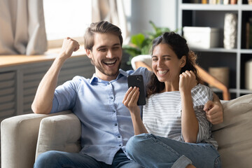 Excited millennial Caucasian couple look at smartphone screen feel euphoric with online win or...