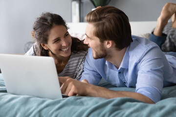Overjoyed millennial couple lying relaxing in bed shopping online on laptop together. Smiling young Caucasian man and woman rest in cozy bedroom at home browse internet on computer.