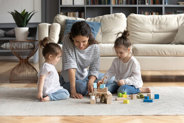 Caring young Caucasian mom sit on floor play with two small daughters construct with building...