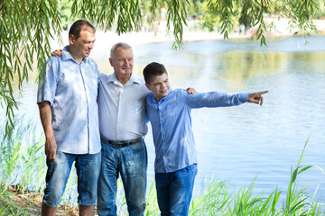 Grandfather, father and son in the summer in nature. Teenager boy points to the right. Three generations of men. Family relationships. Concept of trust. Copy space on the right.